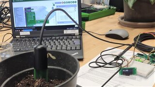Das Foto zeigt einen Topf mit Pflanzenerde, darin steckt ein Sensor, dahinter ein Laptop