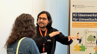 En Mann erklärt einer Frau etwas. Die Frau steht mit dem Rücken zur Kamera. Sie stehen vor einem Banner der KI-Ideenwerkstatt