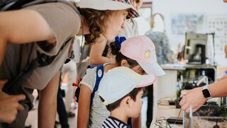 Zwei Kinder und eine erwachsene Person lassen sich von einer Mitarbeiterin eine Versuchsanordnung erklären.