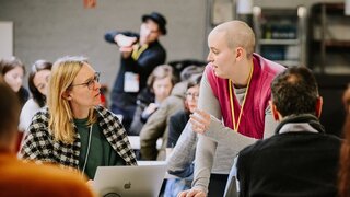 Eine stehende Person erklärt einer sitzenden Frau mit Laptop etwas