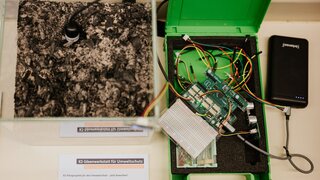 Das Bild zeigt ein Gefäß mit Planzenerde, daneben ein Messgerät, davor eine Postkarte mit dem Motiv der KI-Ideenwerkstatt für Umweltschutz