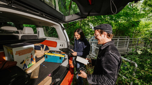 2 Männer stehen an geöffneter Heckklappe von VW-Bus und halten Technik in der Hand