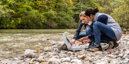 Lowinkelansicht von zwei Forscherinnen, hockend an einem steinigen Flussufer und Messdaten auswertend