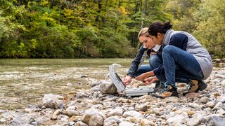 Lowinkelansicht von zwei Forscherinnen, hockend an einem steinigen Flussufer und Messdaten auswertend