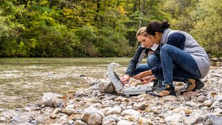 Lowinkelansicht von zwei Forscherinnen, hockend an einem steinigen Flussufer und Messdaten auswertend