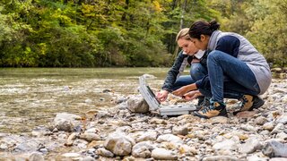 Lowinkelansicht von zwei Forscherinnen, hockend an einem steinigen Flussufer und Messdaten auswertend