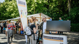 Ein Mann und eine Frau im Gespräch. Neben ihnen steht ein Bildschirm und eine große Flagge der KI Ideenwerkstatt. Im Hintergrund ist ein weiterer Stand zu sehen.