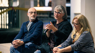 Drei Personen sitzen im Halbkreis. Die mittlere Person spricht in ein Mikrofon.