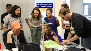 Eine Personengruppe steht um einen Tisch mit Versuchsaufbauten, die der Workshopleiter vorstellt.