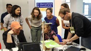 Eine Personengruppe steht um einen Tisch mit Versuchsaufbauten, die der Workshopleiter vorstellt.