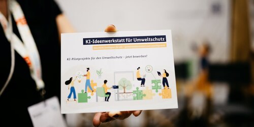Eine Hand hält eine Postkarte mit Informationen über die KI-Ideenwerkstatt dicht vor die Kamera.