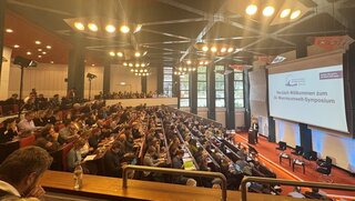 Ein voller Hörsaal, rechts ist eine große Leinwand zu sehen.