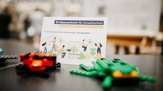 Zwei aus Legobausteinen gebaute Insekten vor einem Flyer der KI-Ideenwerkstatt