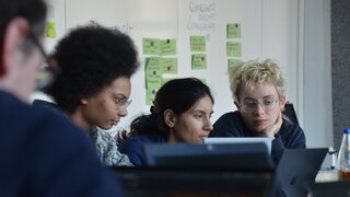 eine Gruppe junger Menschen schaut in Laptops, im Hintergrund Stellwände mit Karten