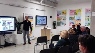 Ein Mann zeigt auf einem Bildschirm mit einer Präsentation, Publikum hört ihm zu