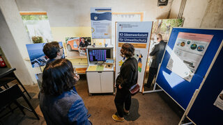 An einem kleinen Messestand sind ein Rechner, Bilder und eine Leinwand mit Plakt zu sehen. Einige Leute stehen davor. 