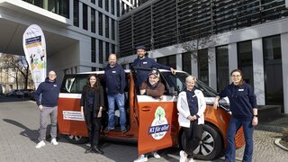 4 Männer und 3 Frauen stehen an einem orangenen E-VW-Bus gelehnt. Sie tragen alle blaue Hoodies mit Logo