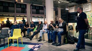 Personen sitzen auf Hockern im Halbkreis. Eine Person steht am Rand. Eine sitzende Person spricht, die anderen hören zu.