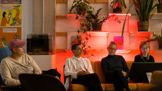 Vier Personen sitzen in einem Halbkreis vor einer Regalwand mit Blumen und unterhalten sich. 