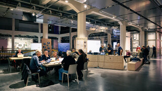 In einer großen Halle sind Tischgruppen aufgebstellt, an denen Personen sitzen und arbeiten 