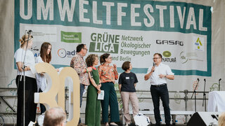 Auf einer großen Bühne stehen mehrere Personen. Der Bundesumweltminister spricht ins Mikrofon. Im Hintergrund hängt ein großes Banner mit der Aufschrift Umweltfestival.