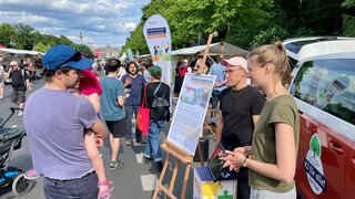 Ein Mann unterhält sich mit einer Frau und einem weiteren Mann vor dem orangenen Bus der KI Ideenwerkstatt. Neben ihnen steht ein Plakat.