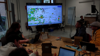Teilnehmende eines Workshops sitzen an einem Tisch mit vielen Laptops, davor ein großer Monitor.