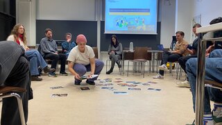 Eine Person mit rosa Mütze hockt in einem Kreis von Personen auf Stühlen und verteilt Bilder auf dem Fußboden. 