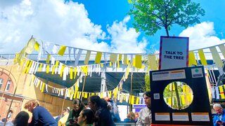 Baum mit Schild "Talk to the Tree", Box mit riesiger Diskette, auf der Anleitung steht, daneben Menschen