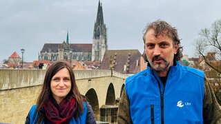 Eine Frau und Mann stehen lächelnd vor der Kulisse einer Brücke und Kirche