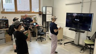 Eine Person steht mit einer VR Brille vor einem großen Bildschirm und wird von einem TV Team gefilmt