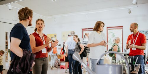 Mehre Personen stehen an einem Messestand und sprechen mit einem Mitarbeiter der KI-Ideenwerkstatt.