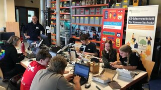 Kinder und Jugendliche sitzen um einen Tisch mit Laptops und bauen eine Vogelbeobachtungsstation. Im Hintergrund sind Regale und Aufsteller der KI IW zu sehen.