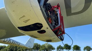 Ein offener Außenlastbehälter sitzt unter einer Tragfläche eines Leichtflugzeugs.