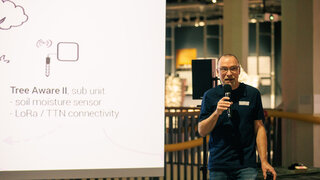 Eine Person steht neben einer großen Leinwand. Er spricht in ein Mikrofon.  