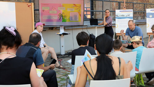 Vor einem Backsteingebäude steht ein Bildschirm, auf dem das Comic zu sehen ist. Davor steht eine Frau an einem Stehtisch und erklärt etwas. Die Zuschauer hören gespannt zu, zu sehen sind ihre Rücken. 