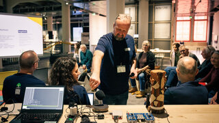 Auf einem Tisch stehen ein Laptop und mehrere kleine Geräte. Ein Mann steht davor und hält ein Mikrofon an einen Lautsprecher auf dem Tisch. Eine Frau sitzt mit dem Rücken zum Betrachter und schaut über die Schulter