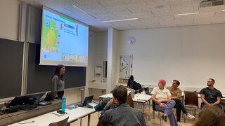 Vor einer großen Leinwand mit der Überschrift "Warum Vogelstimmen" steht eine Person und referiert. Ihr zugewandt sitzen Leute und hören zu. 