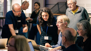 Personen beugen sich im Halbkreis über einen Tisch, an dem Personen sitzen und etwas bauen. 