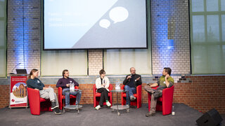 Auf einer Bühne sitzen fünf Personen im Halbkreis auf roten Stühlen. Hinter ihnen ist eine große Leinwand. Sie diskutieren miteinander. 