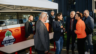 Vor dem orangenen Bus der KI Ideenwerkstatt stehen mehrere Personen und unterhalten sich. Der Bus steht unter einem Pavillon und dient als Registrierung für die Veranstaltung.