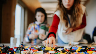 Legosteine, im Hintergrund unscharf zwei Frauen, eine greift nach Steinen